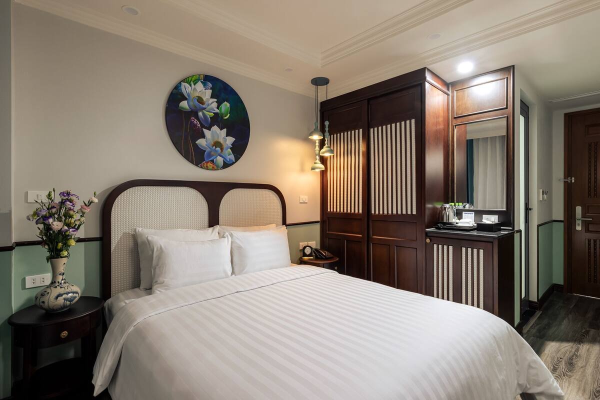 A neatly made bed with white linens, a vase with flowers, a wall-mounted painting, and a wooden wardrobe with a mirror.