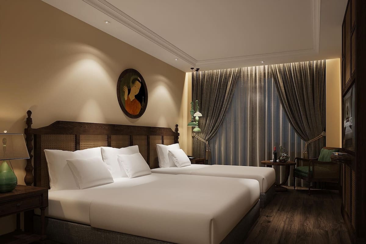 A hotel room with two beds, a wooden headboard, a painting on the wall, a side table with a lamp, and a chair.