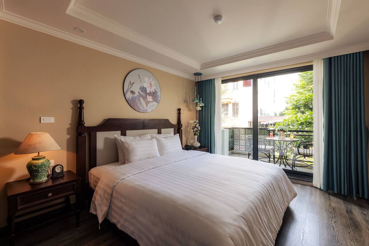 A hotel room with a large bed, a nightstand with a lamp, a wall clock, and a painting. There is a balcony with a view of the outside.