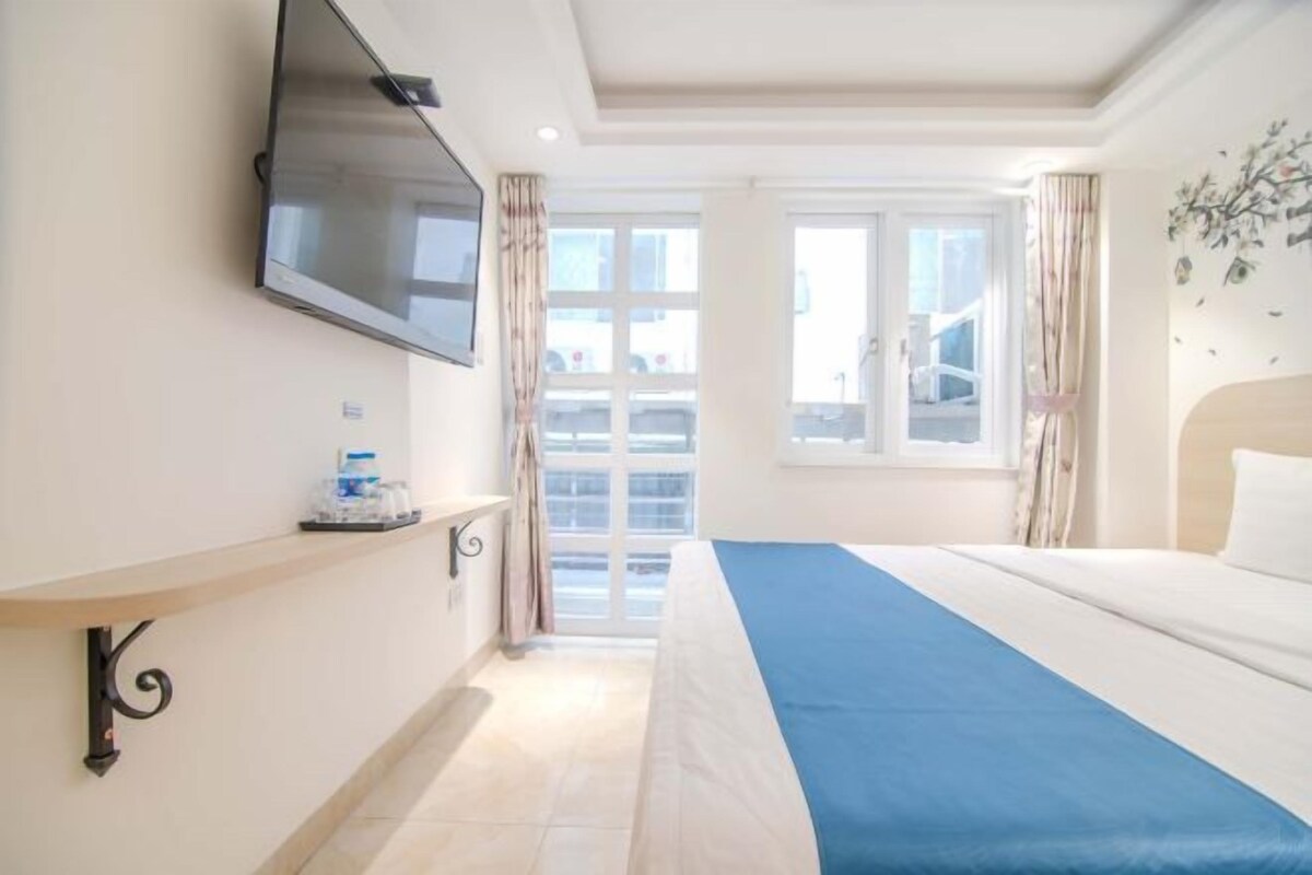 A hotel room with a bed, a TV mounted on the wall, a window with curtains, and a balcony view.