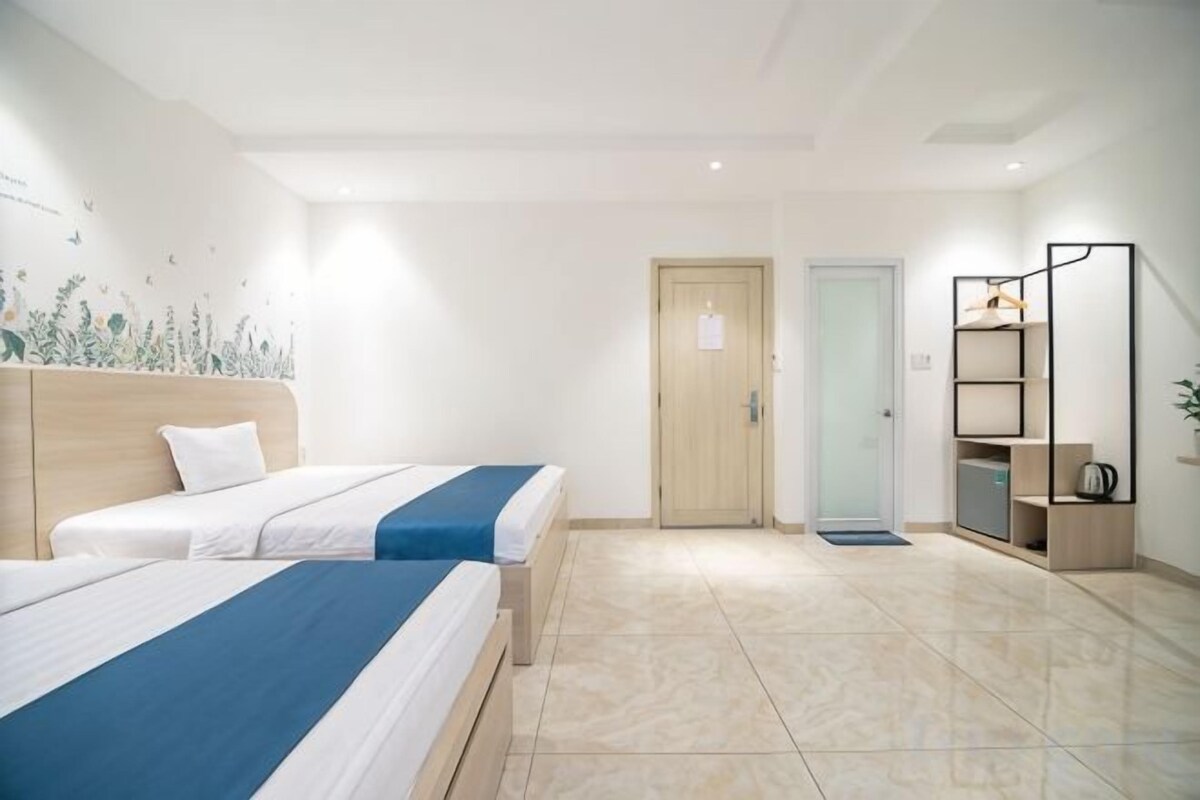 Habitación de hotel con dos camas, cabecero de madera, una puerta, un escritorio pequeño con una silla y una estantería con una planta.