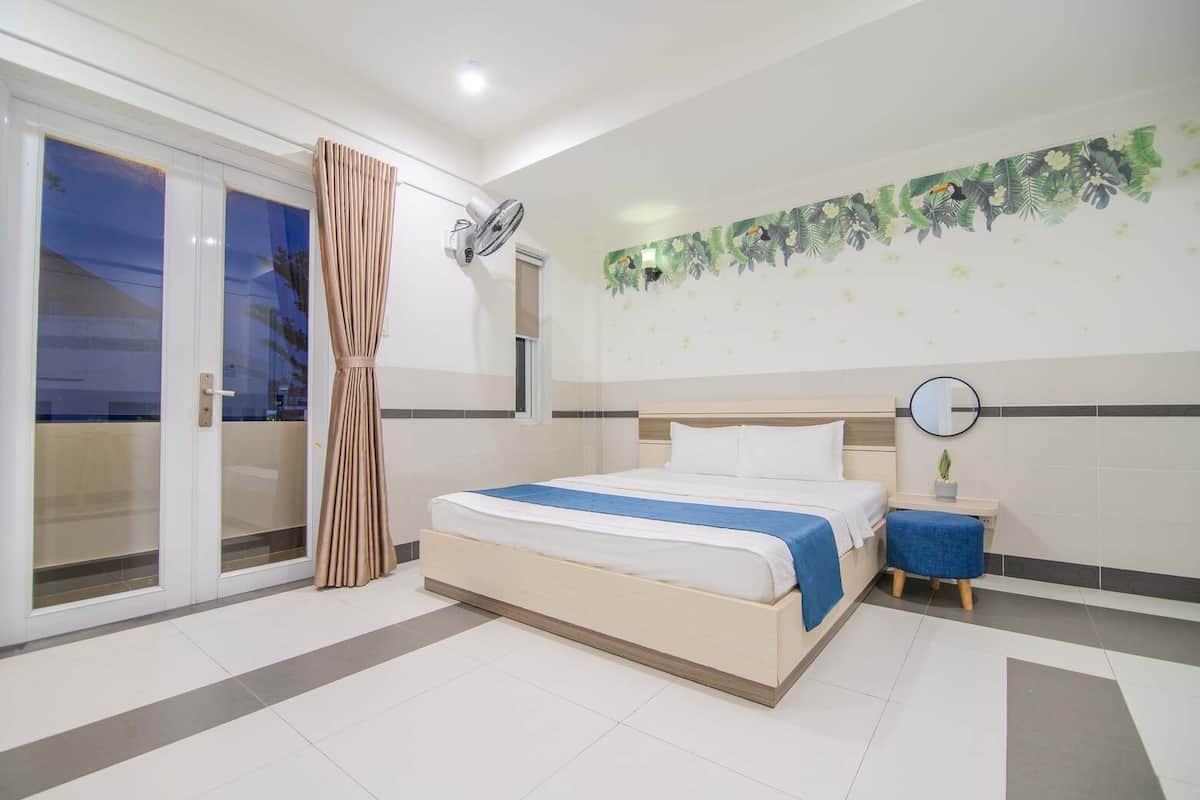 A bedroom with a bed, a nightstand, a mirror, a window with curtains, and a wall with a floral pattern.