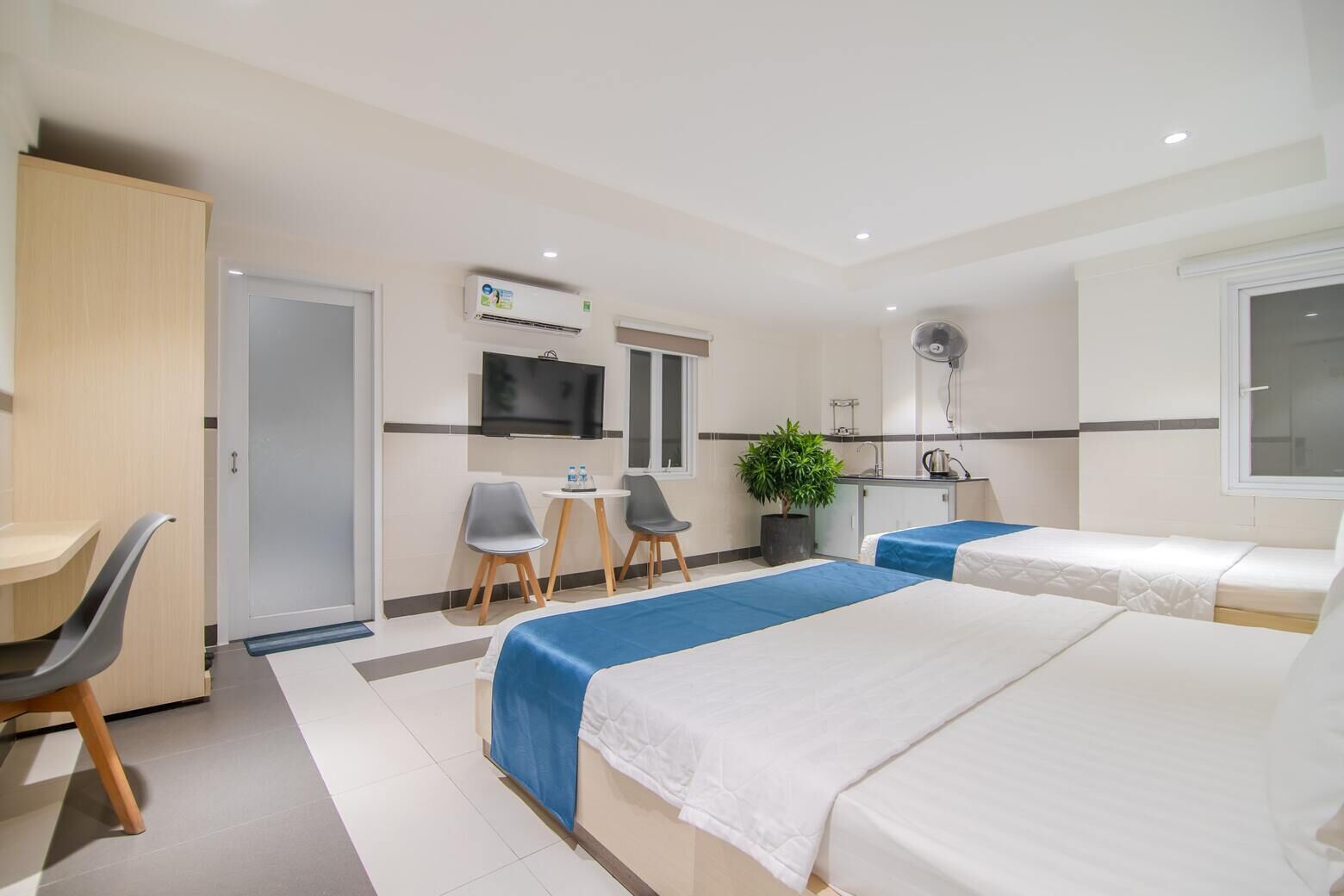 A hotel room with two beds, a desk, a chair, a TV, a small kitchen area, and a potted plant.