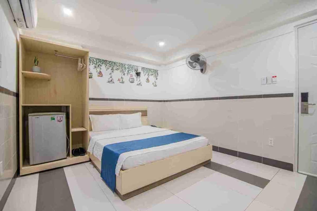 A hotel room with a bed, a small refrigerator, a fan, and a door.