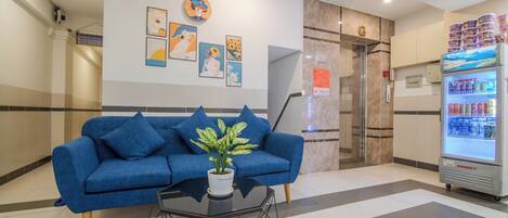 A reception area with a blue sofa, a potted plant, a glass coffee table, a refrigerator stocked with beverages, and a wall with framed artwork.