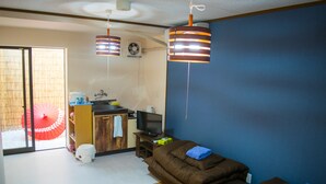 Un espai habitable reduït amb un sofà, una televisió, una cuineta petita i una porta corredissa que dona a un balcó amb un parasol vermell.