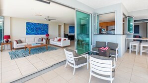 A spacious living area with a white sofa, a coffee table, and two white chairs. A glass door leads to a kitchen area.