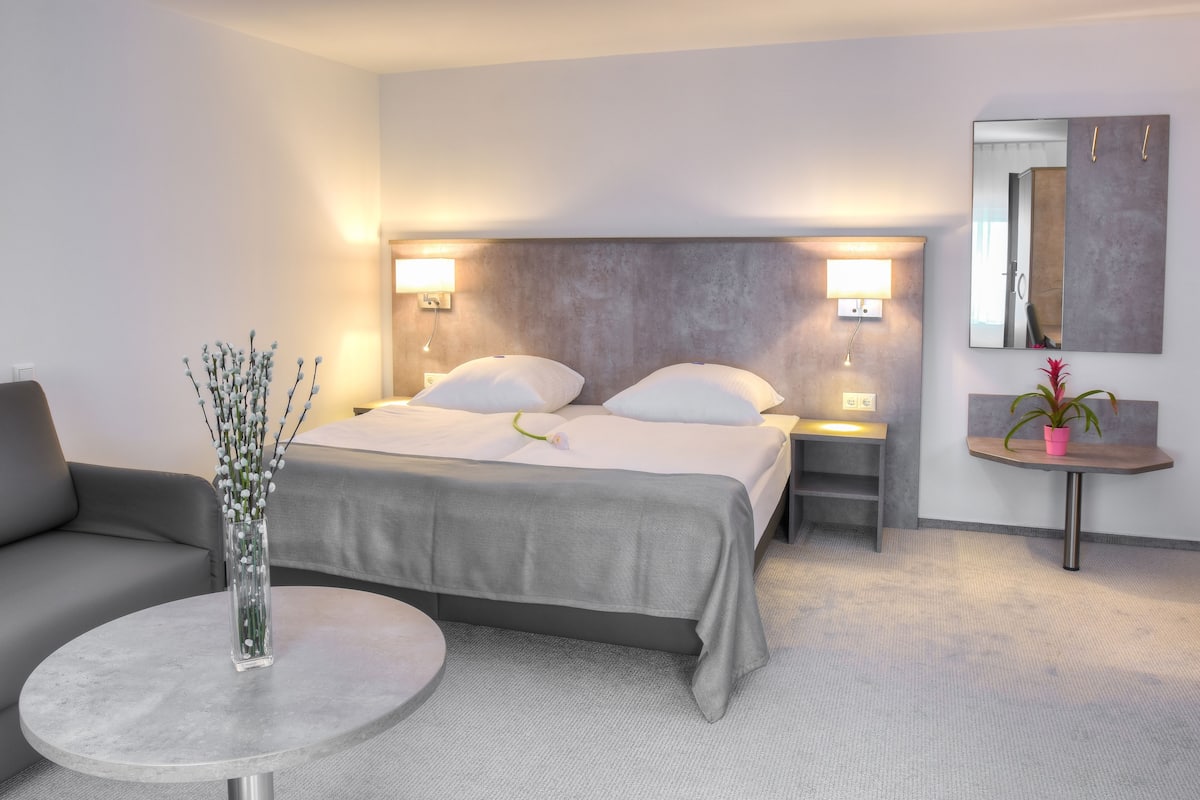 A modern hotel room with a bed, bedside tables, a round table, a sofa, a vase with flowers, and a mirror on the wall.