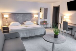 Una habitación de hotel moderna con una cama grande, mesitas de noche con lámparas, una pequeña mesa redonda con una planta, un escritorio con una silla y un televisor montado en la pared.