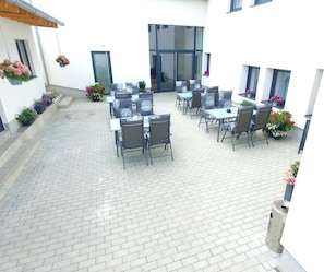 Un patio al aire libre con varias mesas y sillas, rodeado de plantas en macetas y un piso de ladrillo.