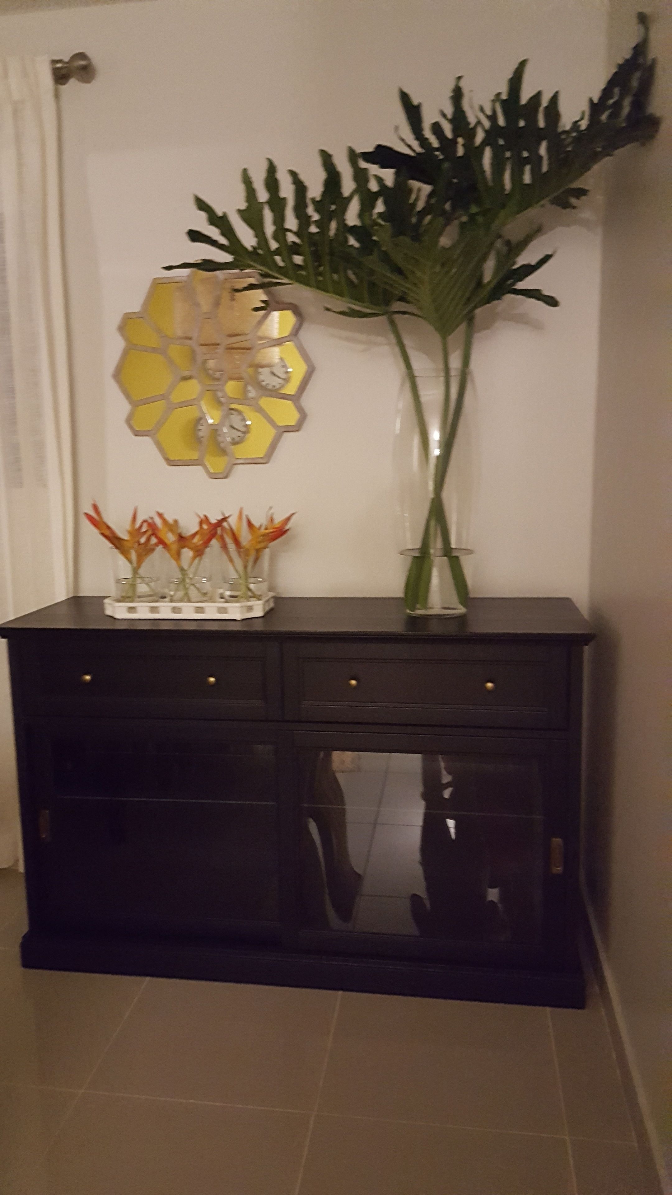A dark wooden cabinet with multiple drawers and a glass door, topped with a decorative yellow hexagonal piece and a tall vase with green foliage.