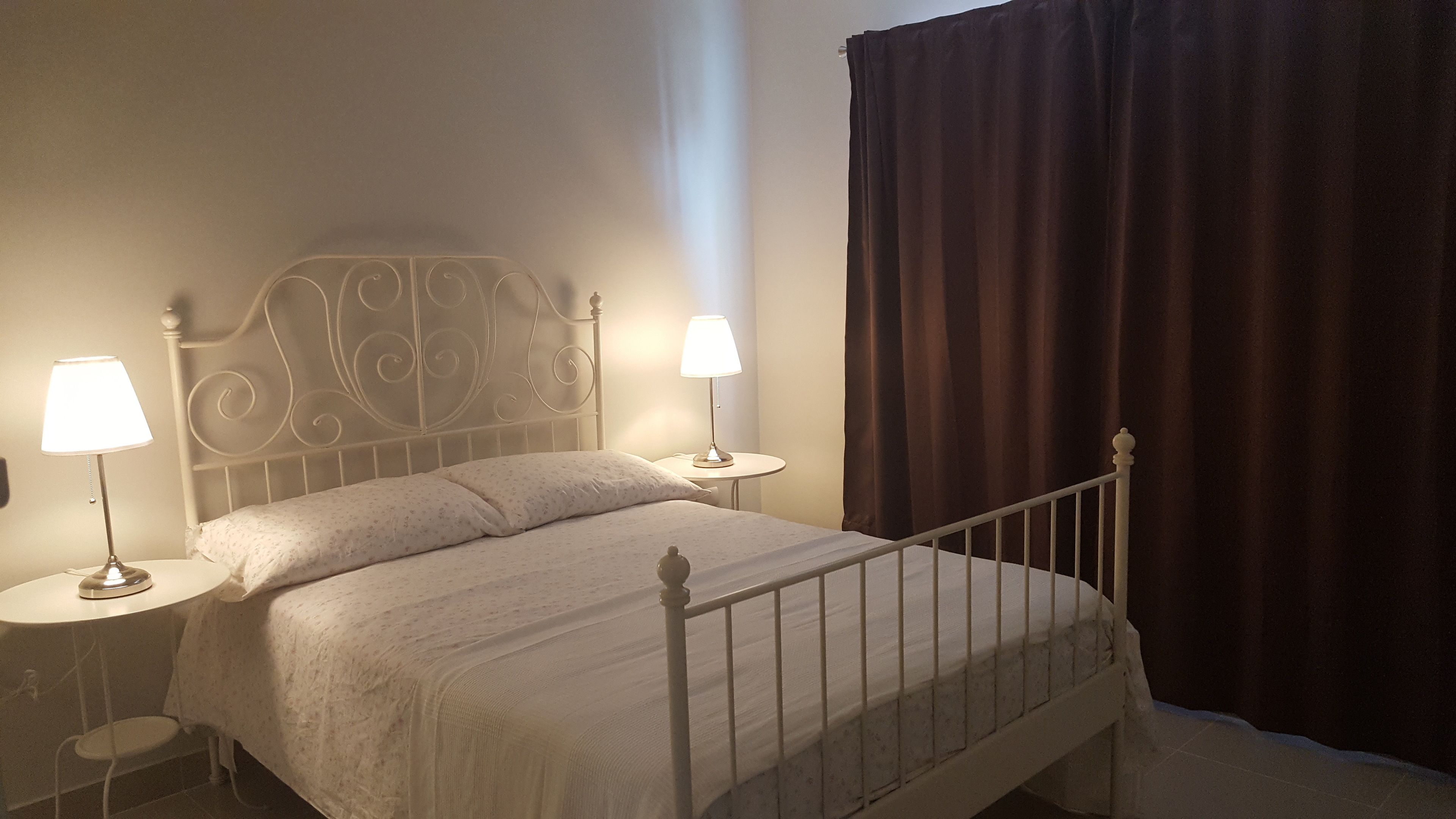 A neatly made bed with a metal headboard, two bedside tables with lamps, and a dark curtain.