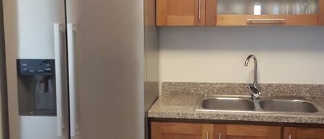 A kitchen with a stainless steel refrigerator, wooden cabinets, a granite countertop, and a stainless steel sink.