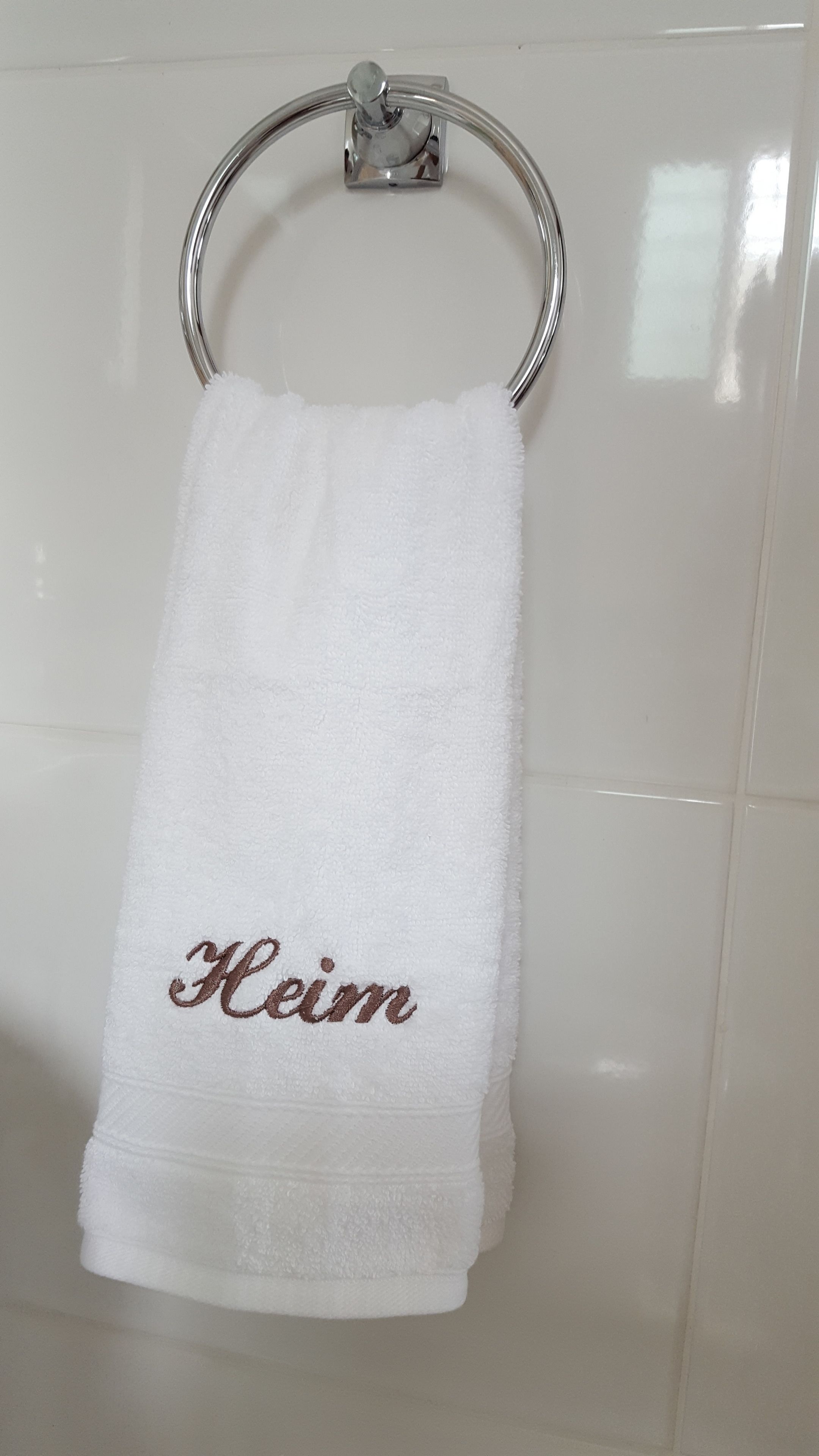 A white towel with the name "Heim" embroidered in brown, hanging on a metal towel ring attached to a tiled wall.