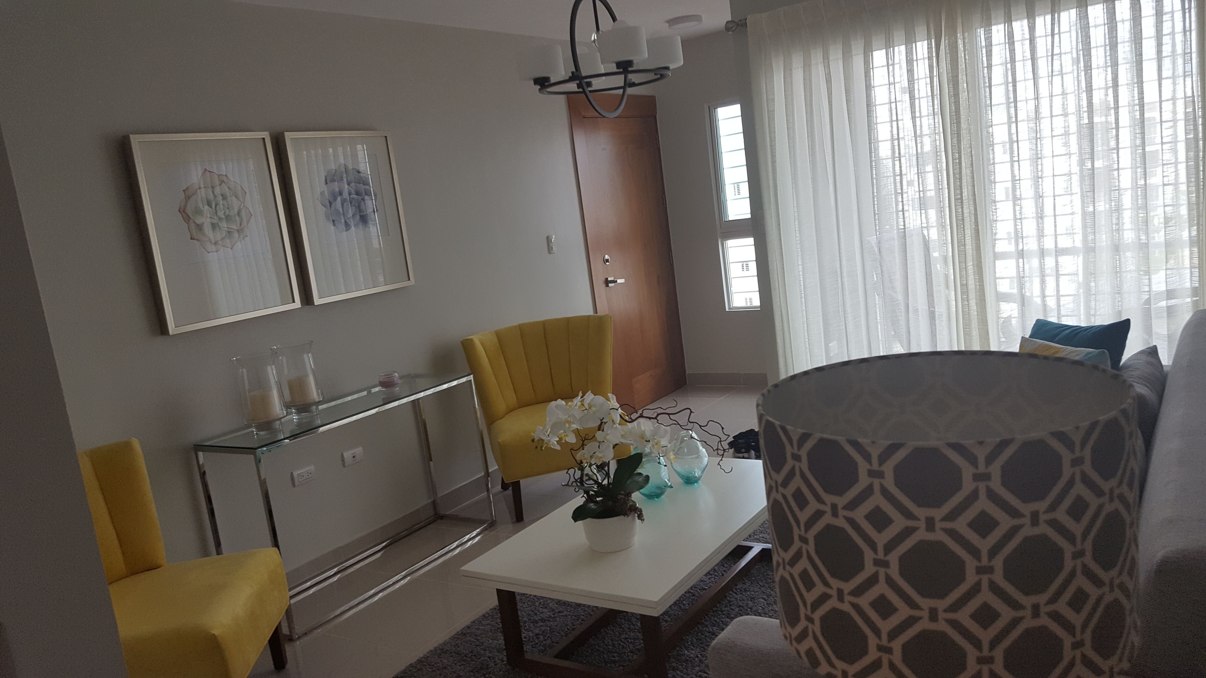 Una sala de estar con un sofá, un sillón amarillo, una mesa de centro de vidrio y un armario blanco. Hay dos cuadros enmarcados en la pared, una lámpara de araña y una ventana con cortinas.
