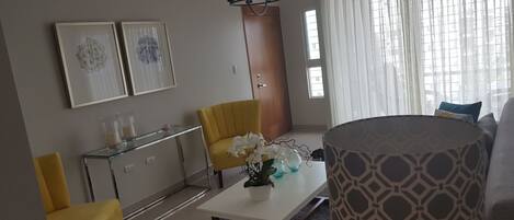 Una sala de estar con un sofá, un sillón amarillo, una mesa de centro de vidrio y un armario blanco. Hay dos cuadros enmarcados en la pared, una lámpara de araña y una ventana con cortinas.