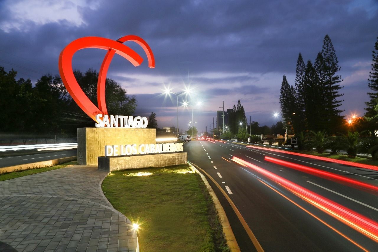 A sign for "Santiago de los Caballeros" with a prominent red sculpture, a paved walkway, and a road with light trails indicating traffic.