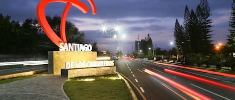A sign for "Santiago de los Caballeros" with a prominent red sculpture, a paved walkway, and a road with light trails indicating traffic.