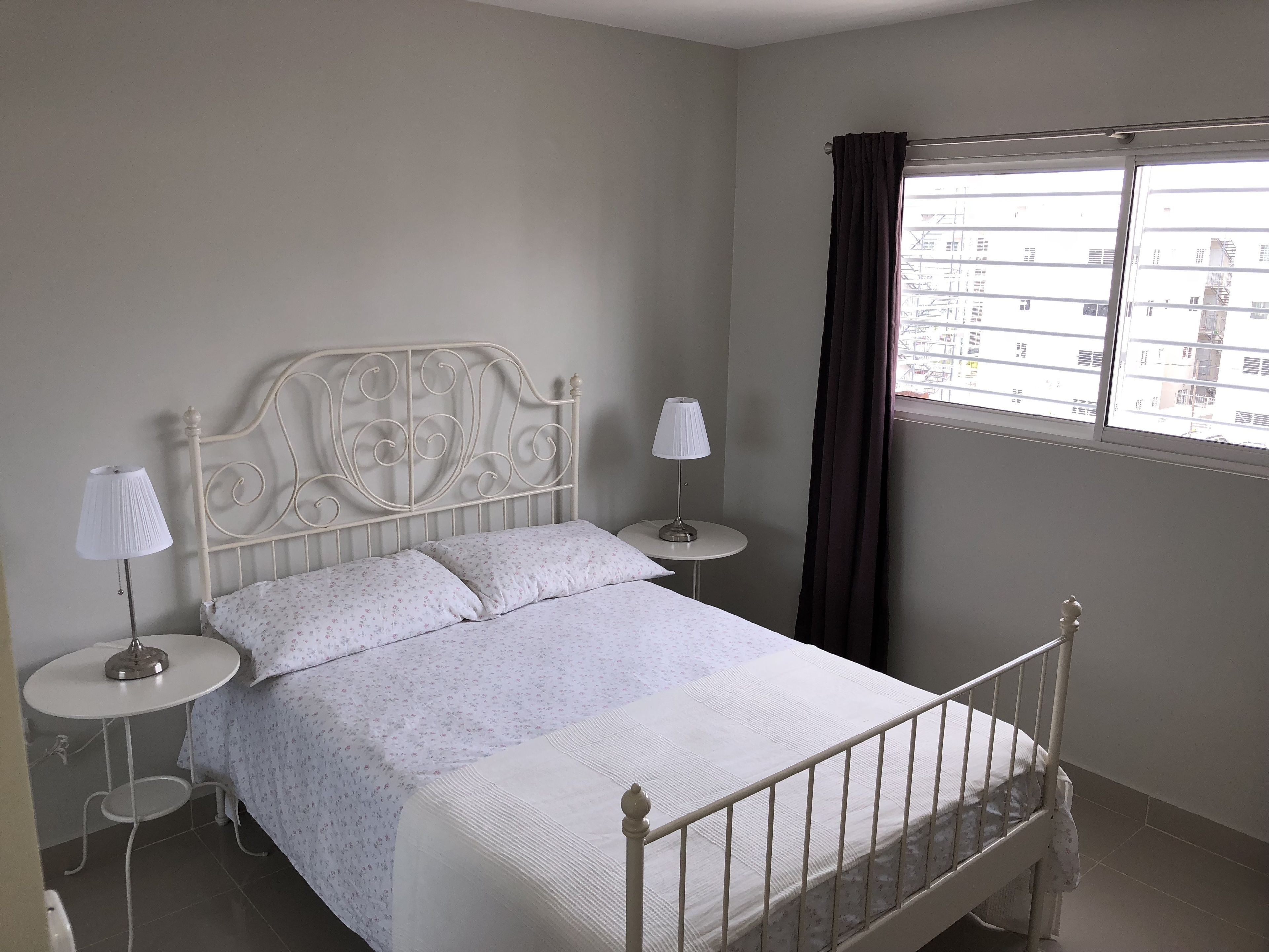 A neatly made bed with a metal headboard, two bedside tables with lamps, a window with curtains, and a view of buildings outside.