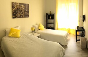 A hotel room with two beds, a small table, a lamp, a shelf with decorative items, and a large window with curtains. - Ficurinia Guest House (Catania)
