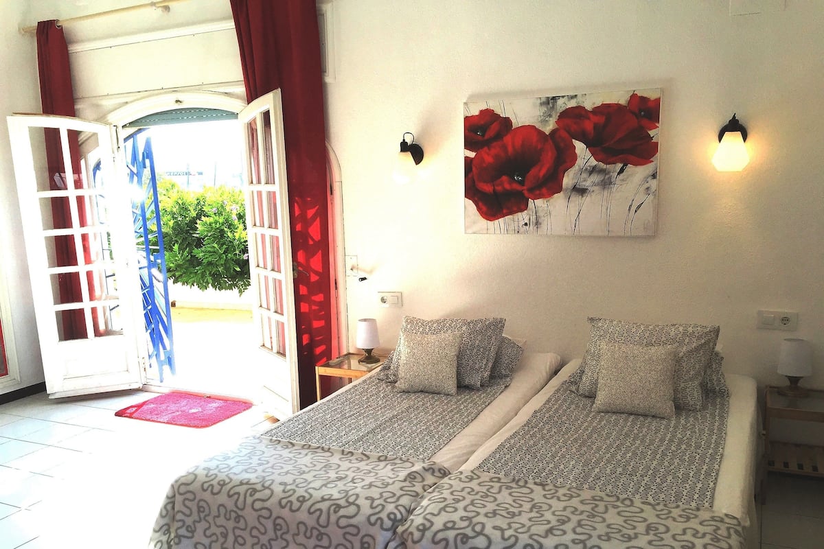 Quarto com cama, pintura de flores vermelhas e vista para o exterior.
