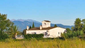Un edificio bianco con un campanile, circondato dal verde e con montagne sullo sfondo.