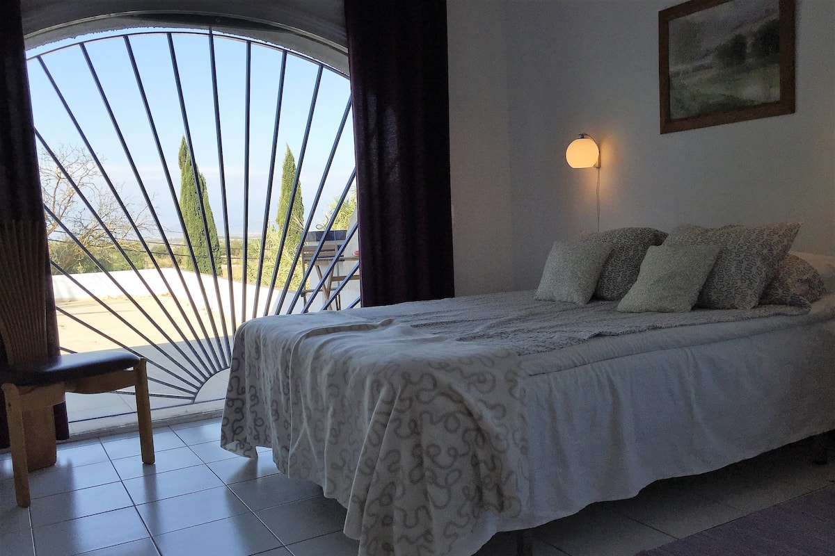 Quarto com cama, cadeira, janela com vista para árvores e luminária na parede.
