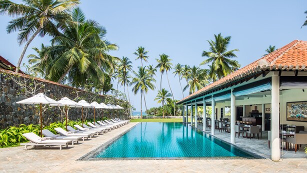 A pool area with lounge chairs and umbrellas, surrounded by palm trees and a building with a tiled roof.