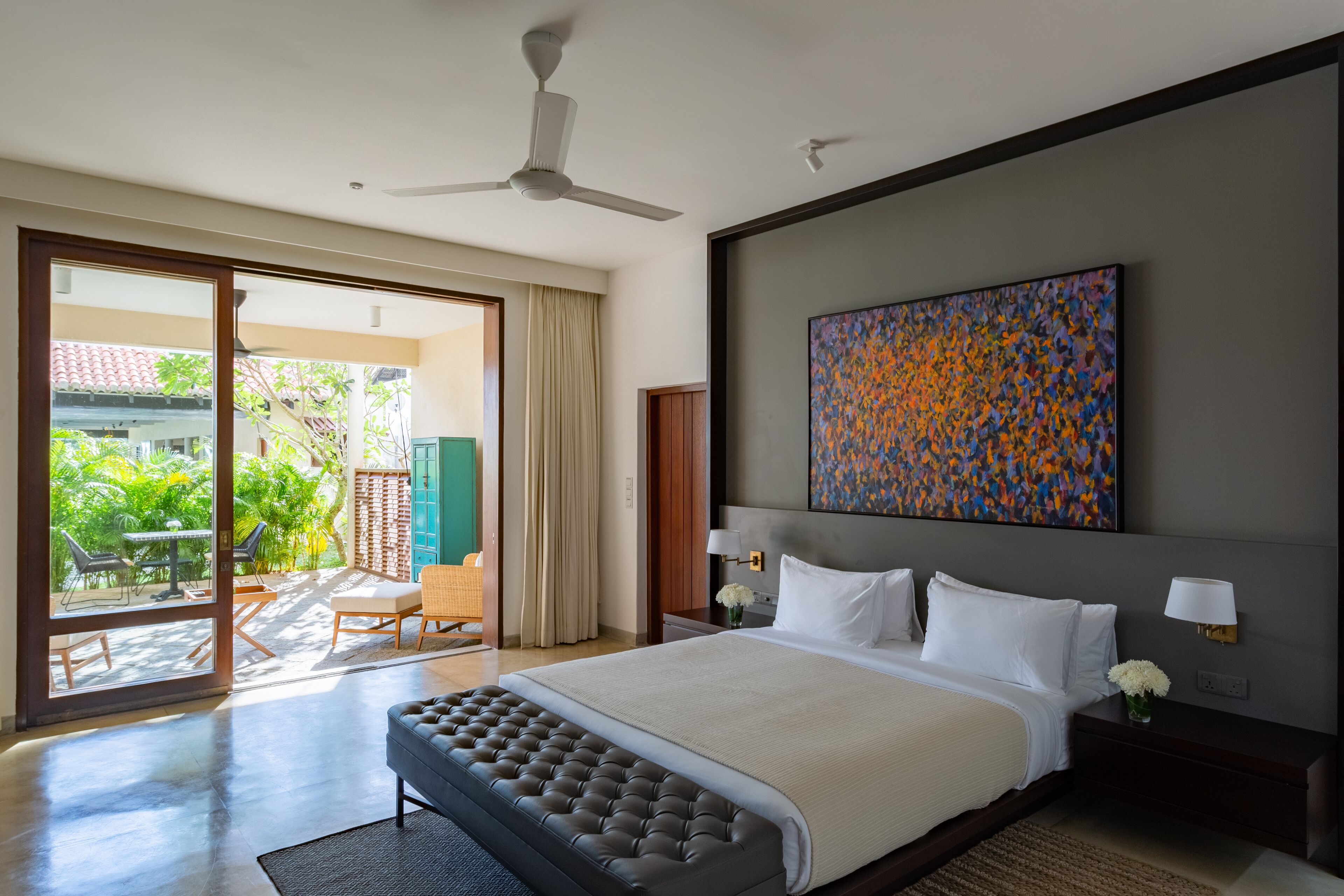 A modern hotel room with a large bed, a ceiling fan, a balcony with outdoor furniture, and a colorful abstract painting on the wall.