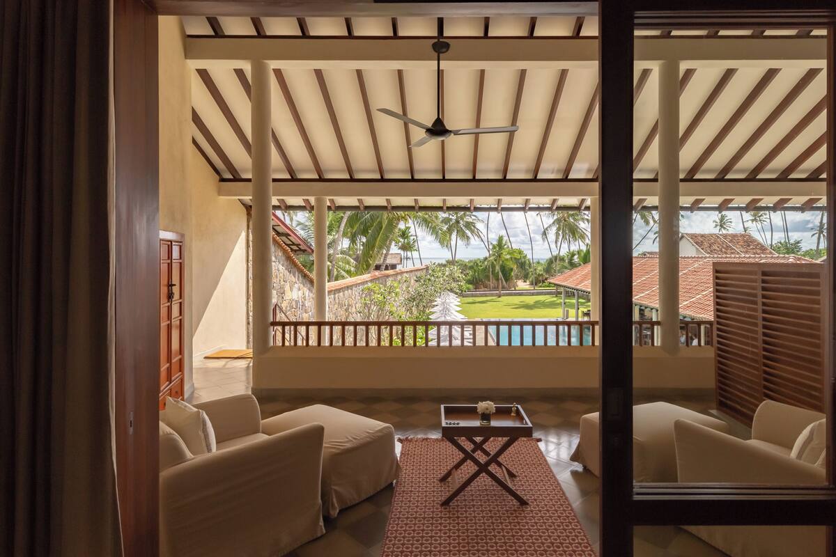 Un área de estar al aire libre techada con ventilador de techo, piscina y vistas a un paisaje tropical.