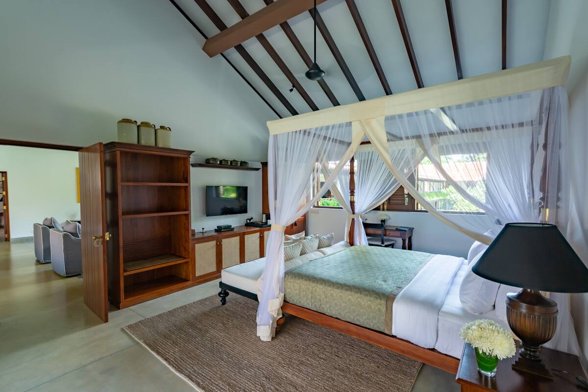 Un dormitorio con una cama con dosel, un televisor sobre un mueble bajo, una estantería de madera y una mesita de noche con lámpara y florero.