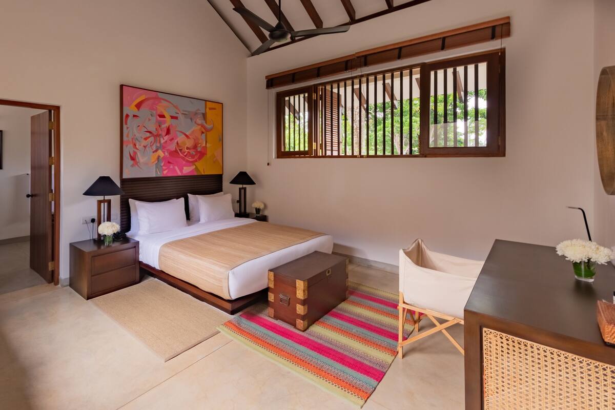 Un dormitorio con cama, mesitas de noche, un escritorio, una silla, una ventana con persianas y un cuadro en la pared.