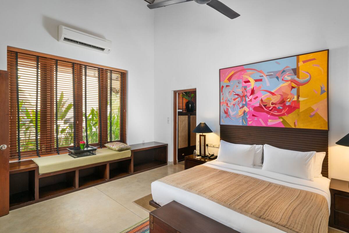 Una habitación de hotel con cama, una banca, una mesita de noche, una lámpara y un cuadro abstracto grande.