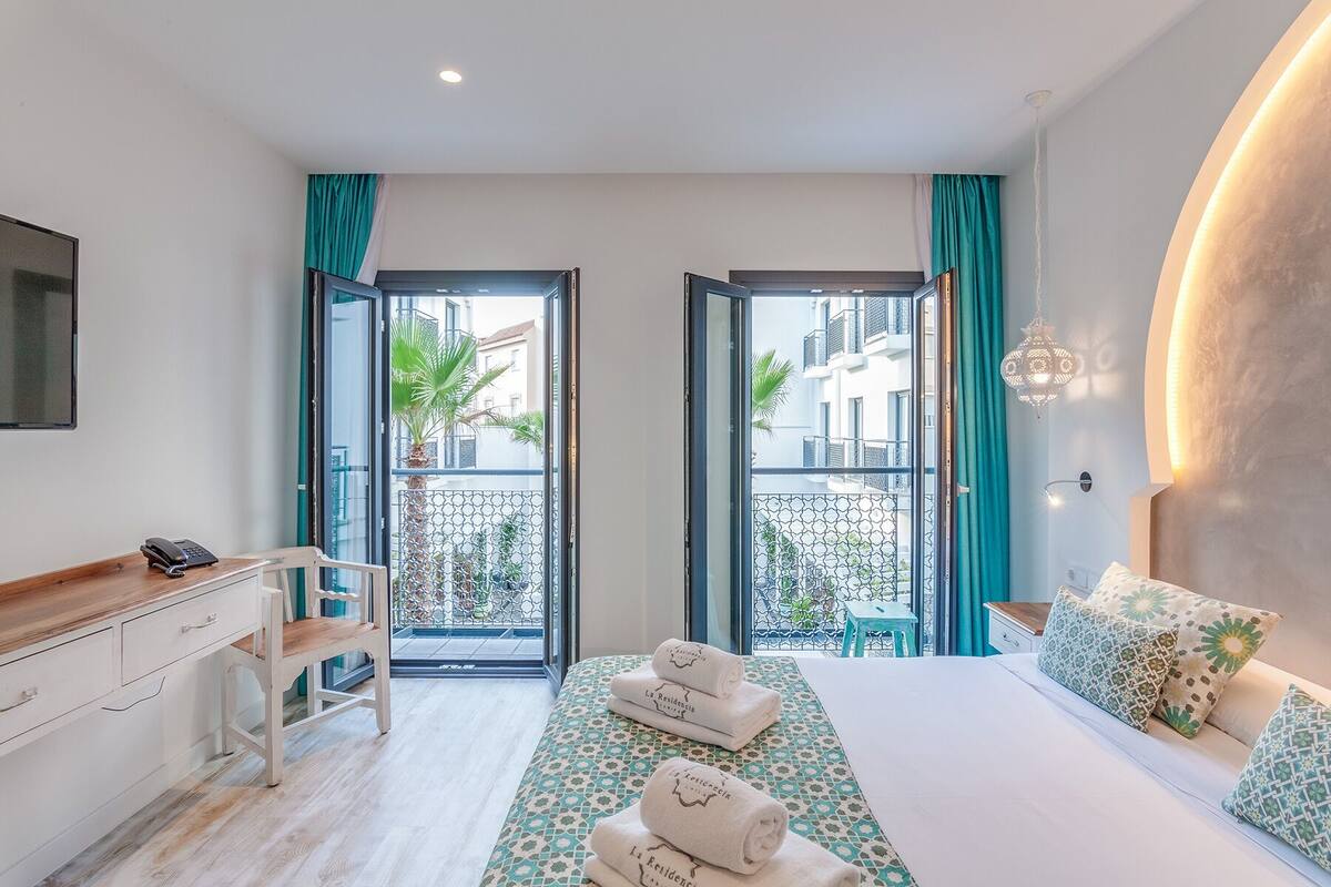Quarto de hotel com cama, escrivaninha com telefone, cadeira, varanda com vista para edifícios e plantas, e luminária pendente.
