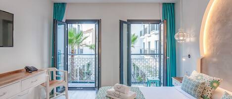 A hotel room with a bed, a desk with a phone, a chair, a balcony with a view of buildings and plants, and a hanging light fixture.