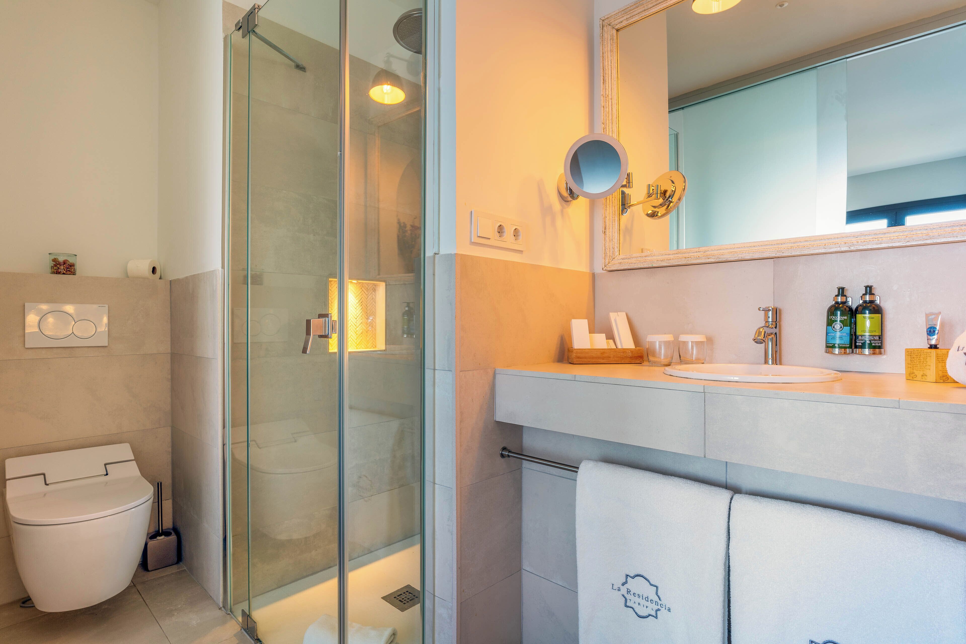 A modern bathroom with a glass shower enclosure, a white toilet, a sink with toiletries, and a mirror with a decorative frame.
