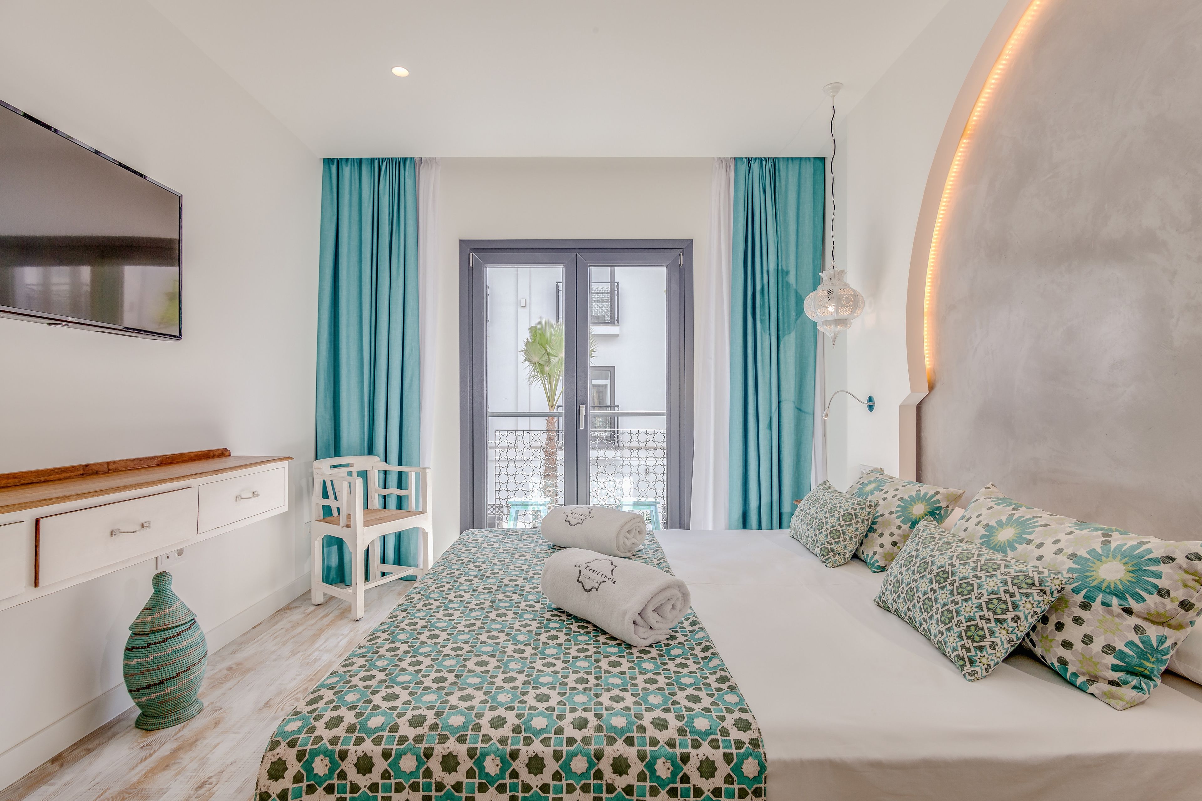A modern bedroom with a patterned bedspread, a white bedside table, a large window with curtains, and a view of the outside balcony.