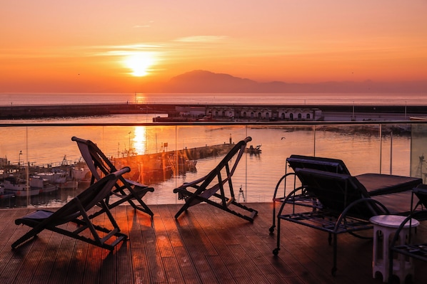A sunset view from a balcony with wooden flooring, two lounge chairs, and a small table.