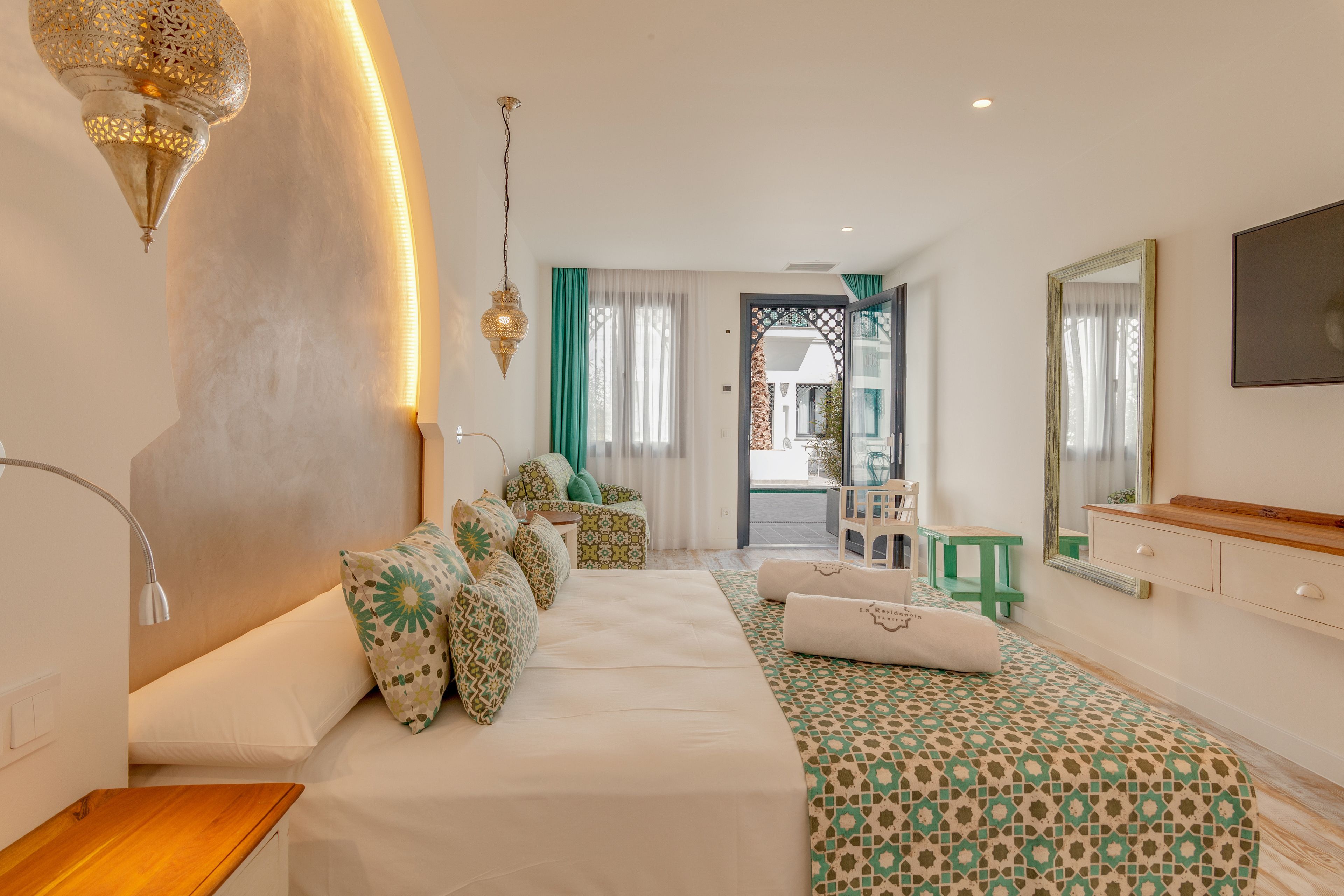 Een moderne hotelkamer met een bank, een bed met een decoratief sprei, een houten nachtkastje, een spiegel en uitzicht op buiten.