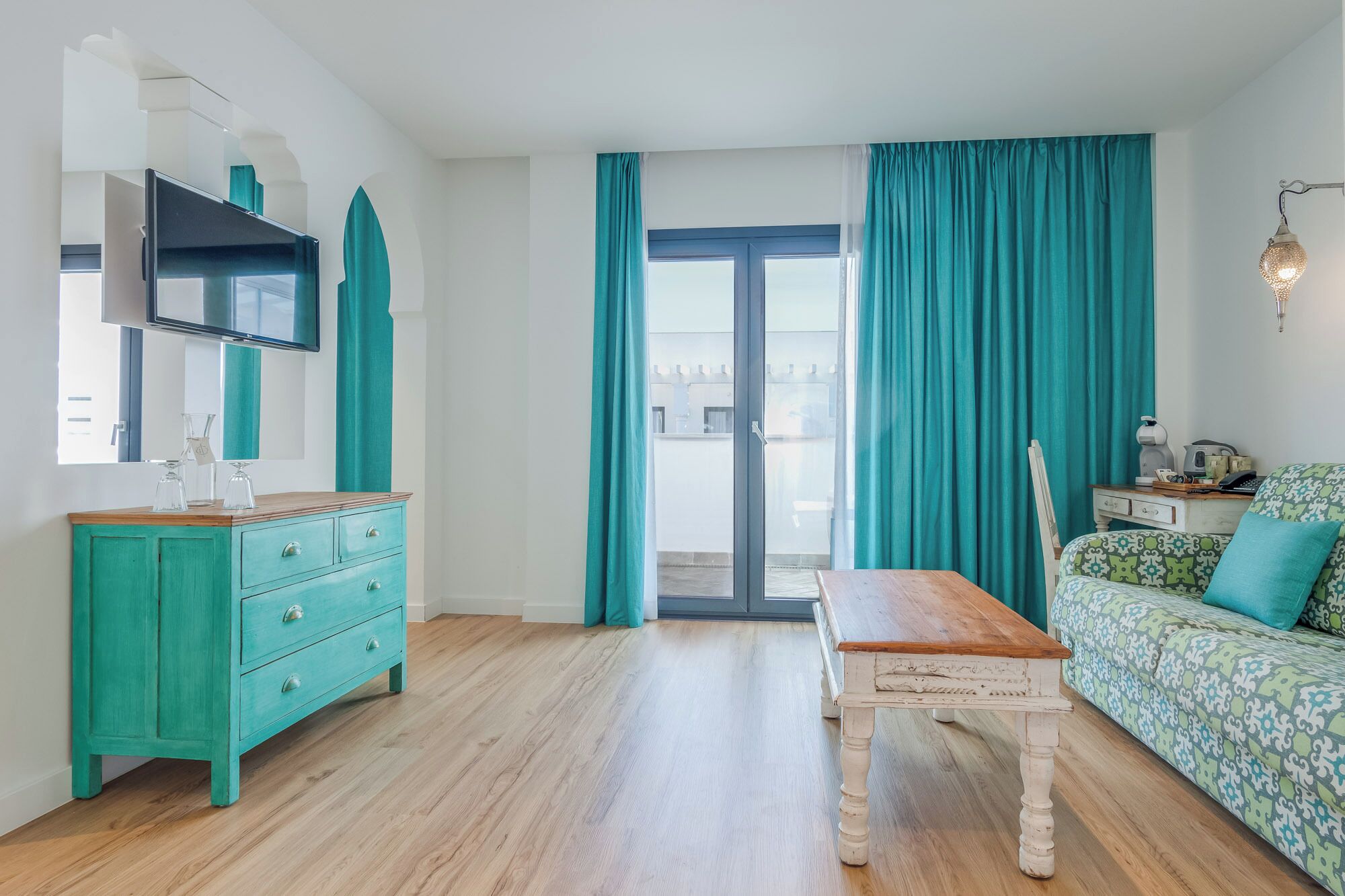 A room with a wooden table, a teal-colored dresser, a sofa with patterned upholstery, and a large window with teal curtains.