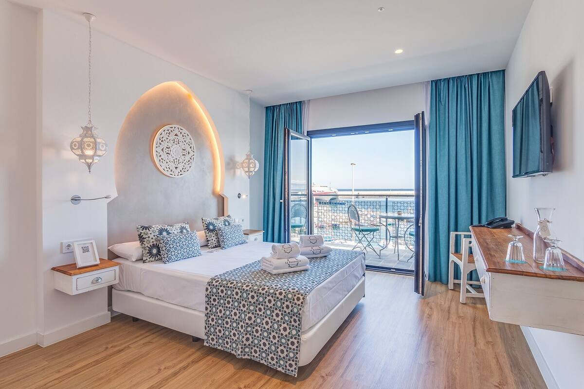 Quarto de hotel moderno com cama, mesas de cabeceira, escrivaninha e varanda com vista para o mar.