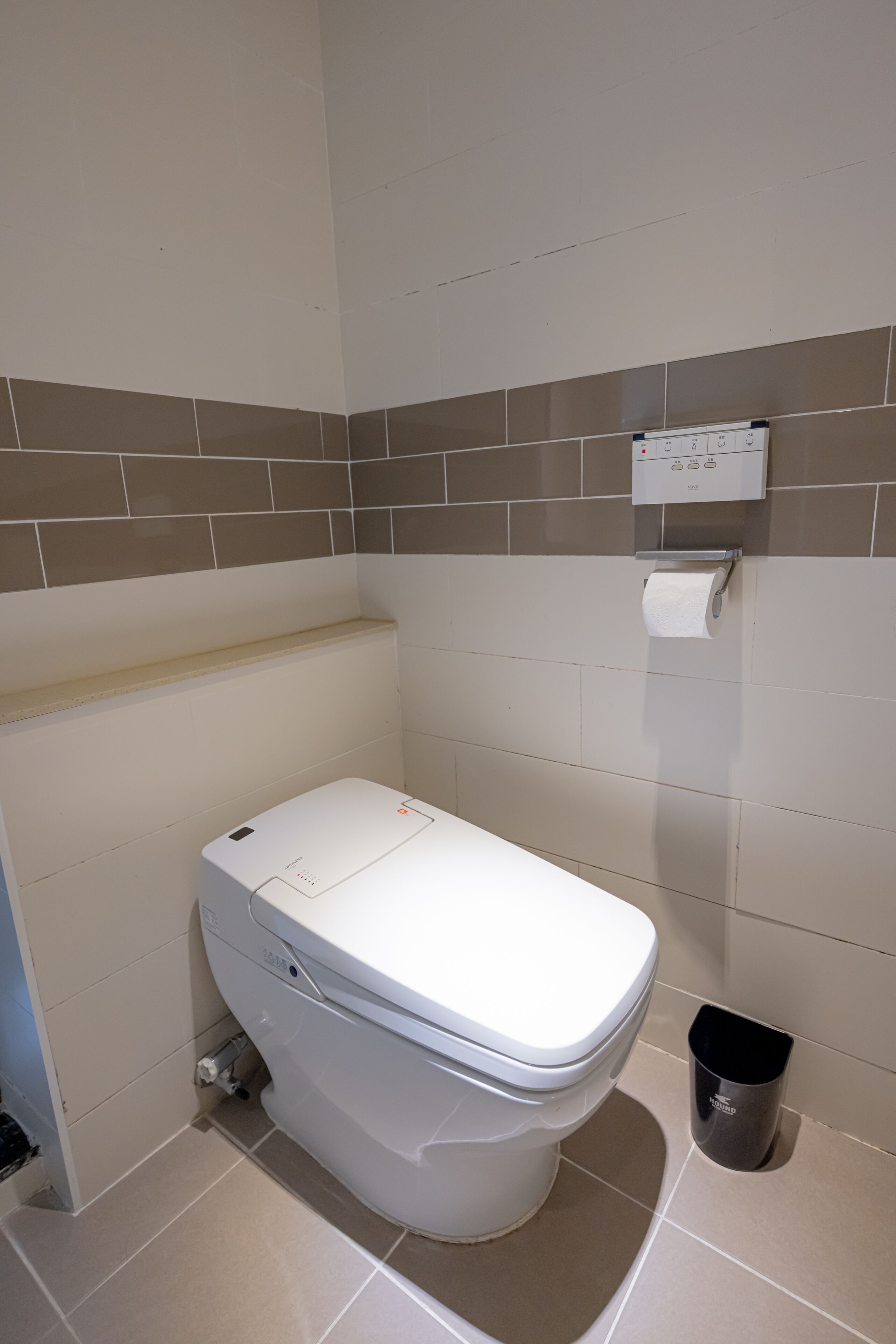 A white toilet with a paper towel dispenser and a trash bin in a bathroom with beige and brown tiled walls.