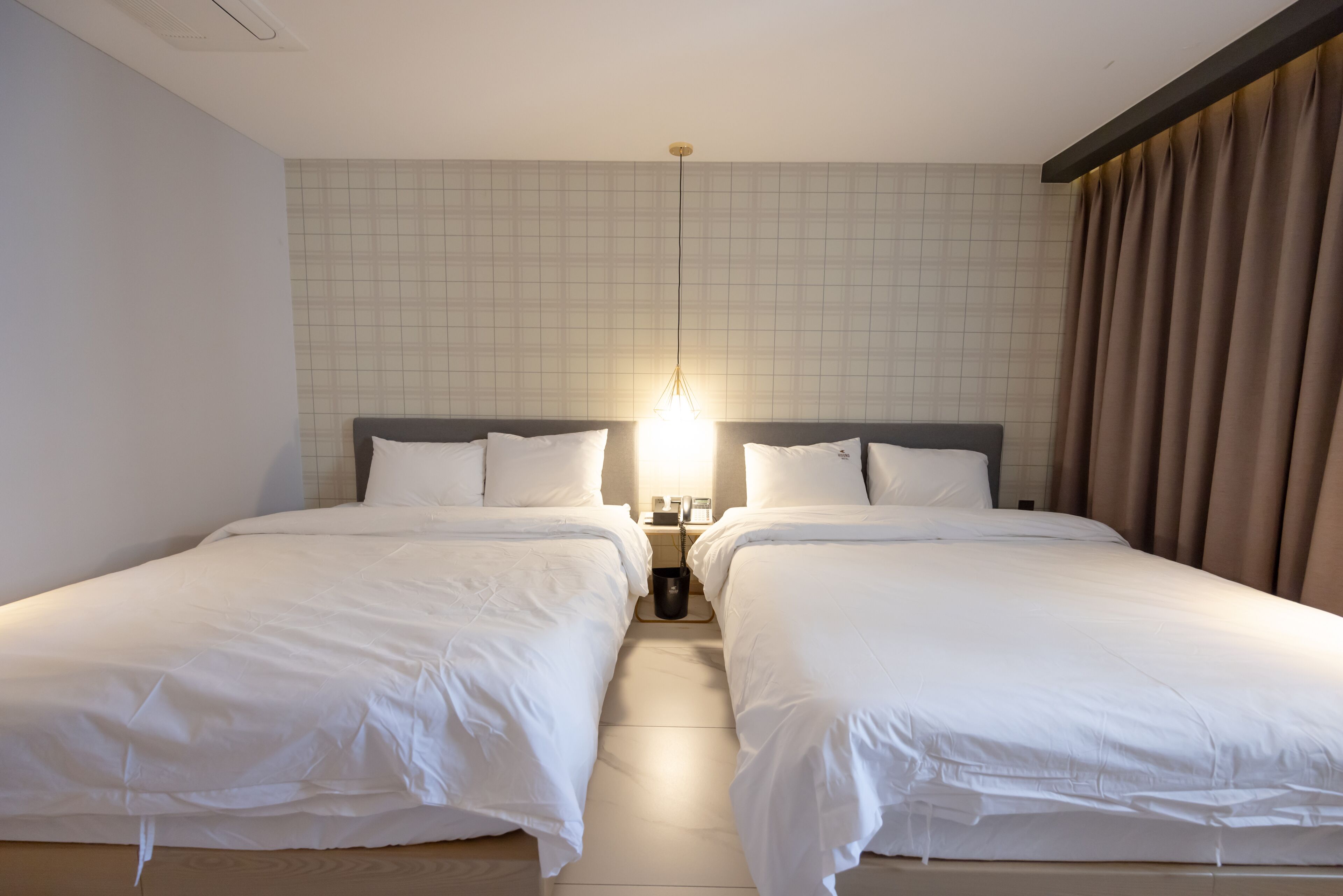 Habitación de hotel con dos camas individuales, una mesita de noche con lámpara y pared de azulejos.