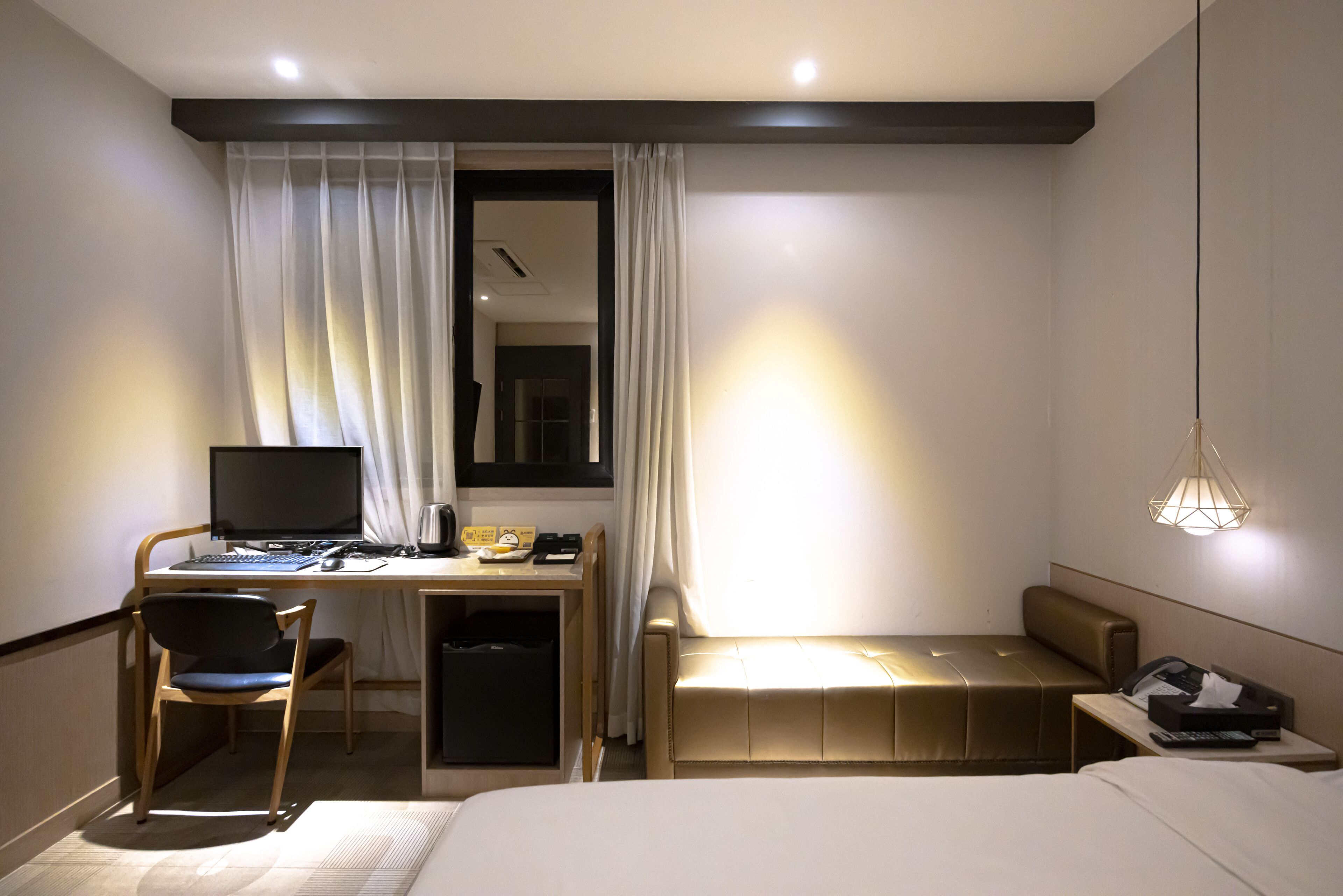 Habitación de hotel con cama, escritorio con computadora, silla, sofá pequeño, mesita de noche con teléfono y lámpara colgante.