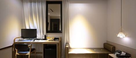 Habitación de hotel con cama, escritorio con computadora, silla, sofá pequeño, mesita de noche con teléfono y lámpara colgante.