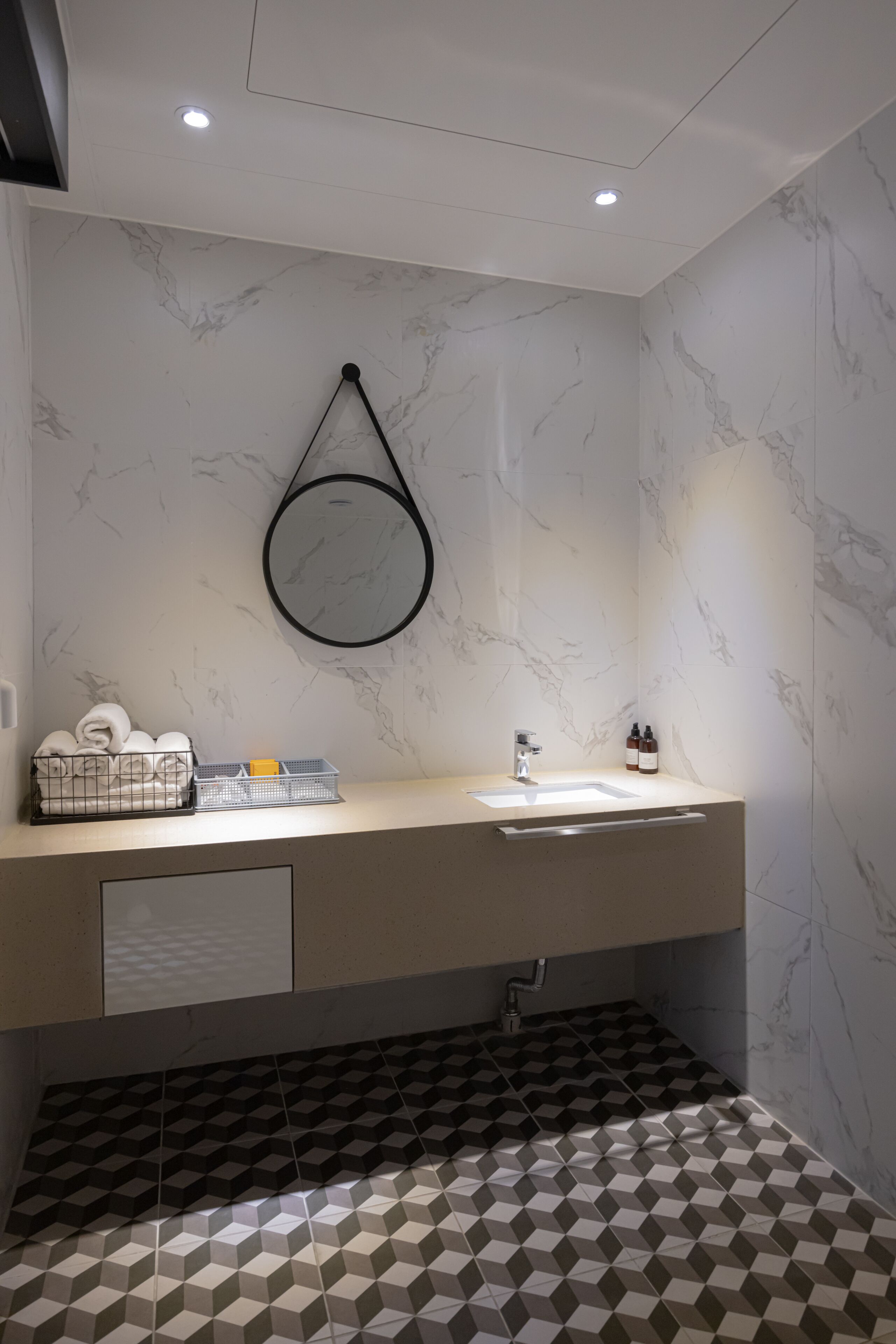 Ein modernes Badezimmer mit Marmorwand, rundem Spiegel, Waschbecken mit Wasserhahn und Fliesenboden.