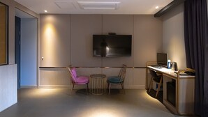 A modern hotel room with a flat-screen TV mounted on the wall, a small round table with two chairs, a desk with a computer and a coffee maker, and a door leading to another room. - Hound Hotel Seomyeon - Bumcheon (Busan)
