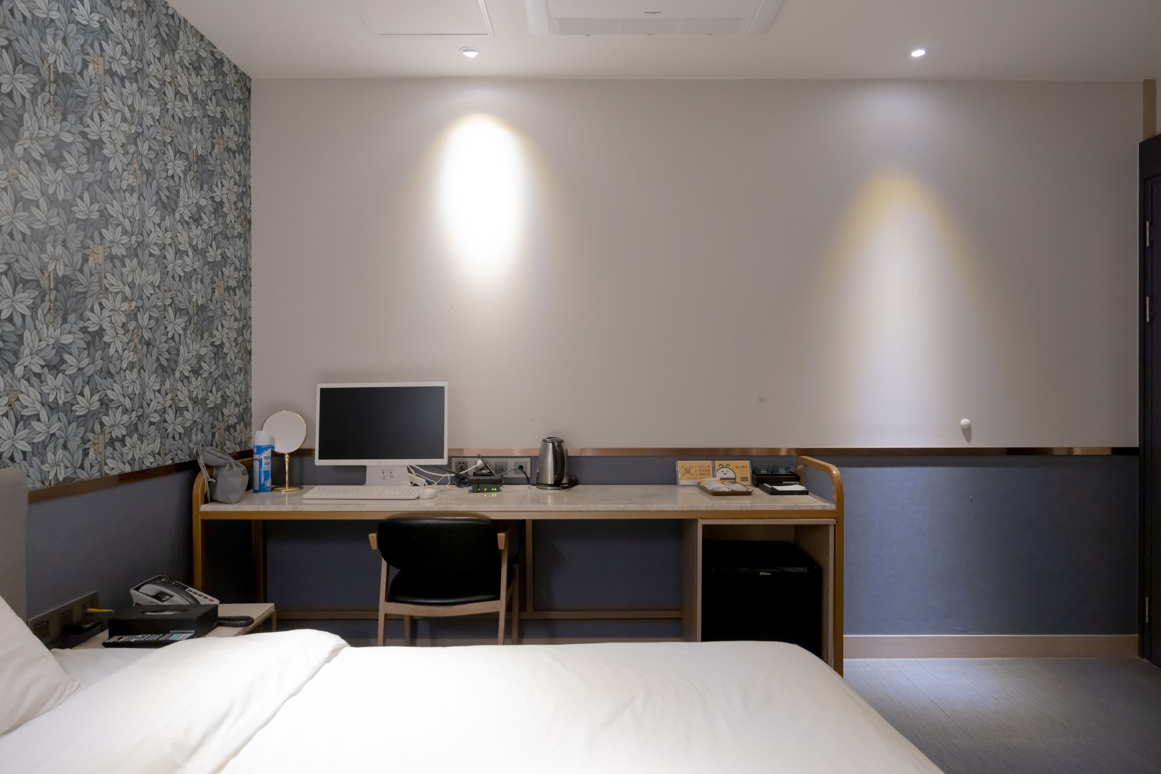 A hotel room with a bed, a desk with a computer, a chair, and a small kitchenette area.