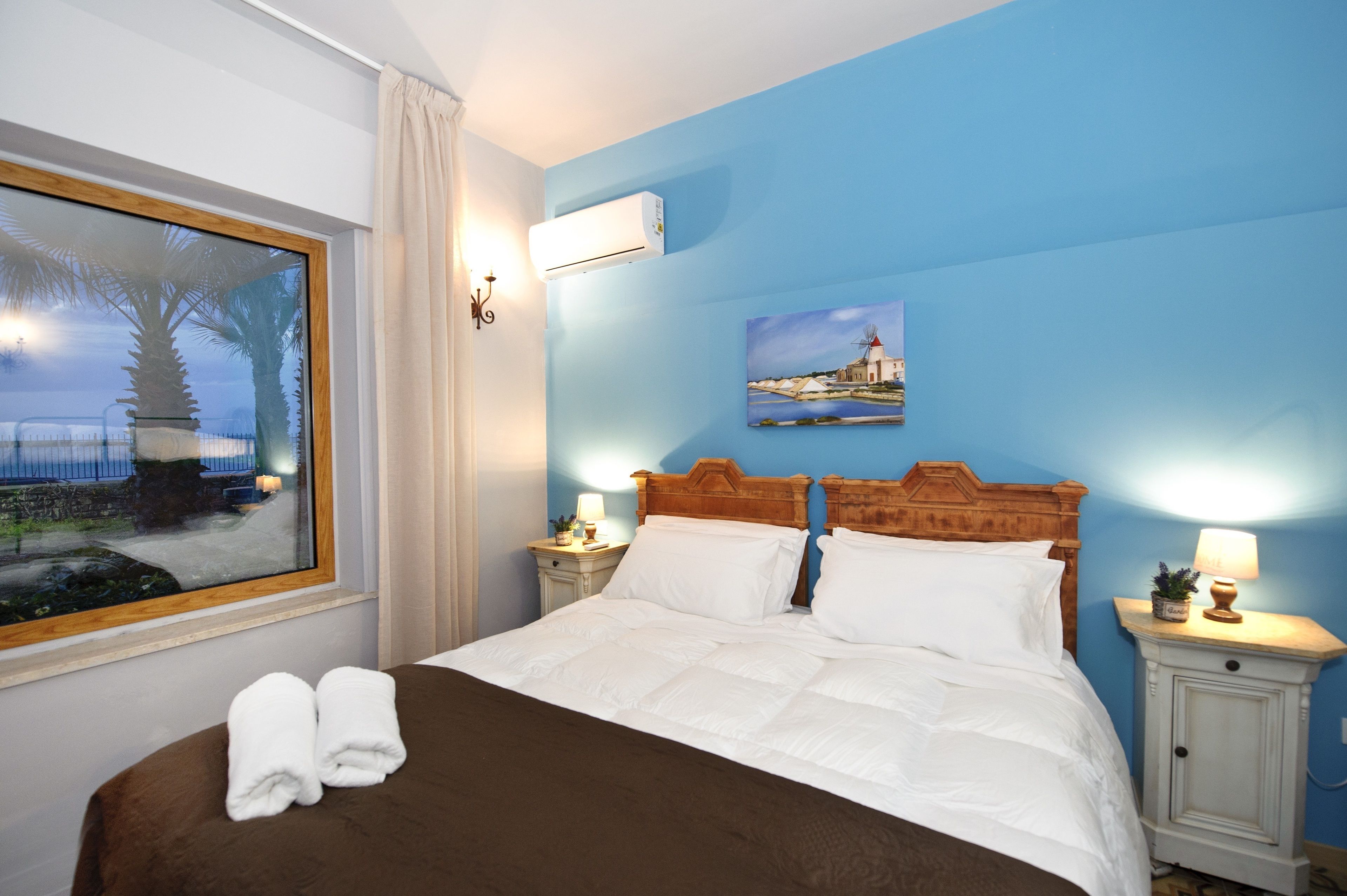 Un cuarto de hotel con una cama grande, dos mesitas de noche con lámparas, un cuadro en la pared y una ventana que ofrece vistas a palmeras y un edificio.