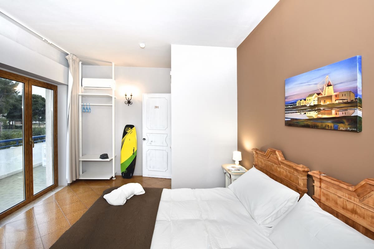 Una habitación de hotel con una cama, una mesita de noche, un cuadro, una puerta y una ventana con vistas al exterior.